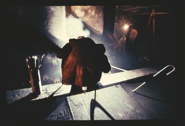 Descrição da imagem: Fotografia horizontal colorida de um trabalhador em um ambiente industrial, visto de costas. O trabalhador usa um casaco grosso e está curvado sobre uma superfície de metal, com vapor ou fumaça subindo ao seu redor. Luzes fortes iluminam parcialmente a cena, destacando o ambiente de trabalho pesado e áspero. Fim da descrição da imagem.