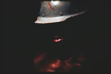 Descrição da imagem: Fotografia horizontal colorida com um enquadramento vertical de uma pessoa com capacete, parcialmente imerso na escuridão. A iluminação destaca apenas parte do rosto e do capacete, criando um forte contraste entre as áreas iluminadas e sombreadas. O capacete de metal tem uma aparência desgastada e apresenta um pequeno desenho de raio vermelho. A pessoa é negra, e a iluminação destaca parte dos lábios e do torso nu. Fim da descrição da imagem.