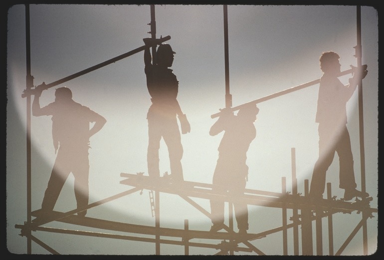 Descrição da imagem: Fotografia horizontal colorida de quatro trabalhadores em um andaime, capturada contra um fundo claro e brilhante. As figuras aparecem em silhueta, destacando seus contornos enquanto manipulam barras de metal. Fim da descrição da imagem.