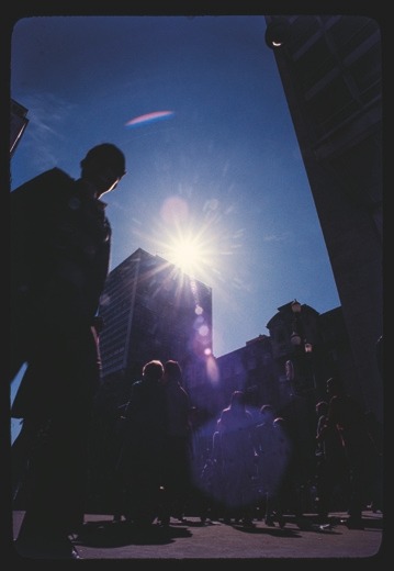 Descrição da imagem: Fotografia vertical colorida de uma cena urbana ao ar livre, onde a luz do sol brilha intensamente no topo de um edifício alto. A cena é vista contra o céu azul, com várias pessoas andando e outras paradas, criando sombras longas. A luz do sol cria um efeito com reflexos coloridos, e os prédios ao fundo estão parcialmente encobertos por sombras. Fim da descrição da imagem.
