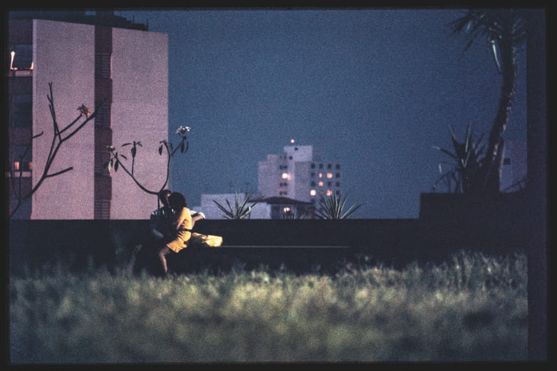 Descrição da imagem: Fotografia horizontal colorida de um casal sentado em um banco de praça a noite. Cercado por alguns prédios altos ao fundo e árvores que aparecem nas laterais da imagem. O casal está em um momento íntimo, sentados um de frente ao outro com iluminação suave vinda de fontes distantes. O gramado em primeiro plano está desfocado. Fim da descrição da imagem.