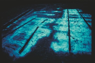 Descrição da imagem: Fotografia horizontal colorida de uma superfície de pavimento rachado, iluminada por uma luz azulada intensa. A imagem mostra sombras e padrões geométricos formados pela iluminação, criando um efeito abstrato. As rachaduras e marcas no solo sugerem um ambiente antigo ou desgastado. Fim da descrição da imagem.