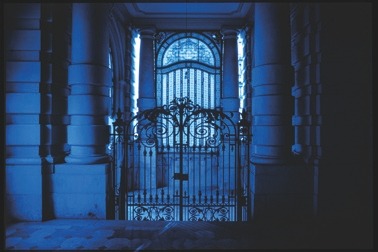 Descrição da imagem: Fotografia horizontal colorida de uma portaria ou entrada de um prédio, iluminada por uma luz azulada. A cena mostra um portão de ferro forjado com detalhes ornamentados, entre duas grandes colunas de pedra. Ao fundo, uma janela arqueada com vidro fosco. Fim da descrição da imagem.