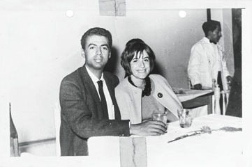 Descrição da imagem: Fotografia em preto e branco de George Love, à esquerda, e Claudia Andujar, à direita, sentados em uma mesa. Ele usa terno e gravata e segura um copo na mão direita. Ela usa um casaco claro por cima do vestido e um colar. Ao fundo, desfocado, um homem negro de perfil direito e usa roupa social branca. Fim da descrição da imagem.