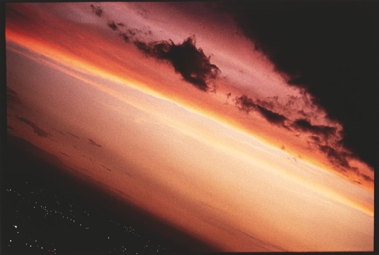 Descrição da imagem: Fotografia horizontal colorida de um pôr do sol. A imagem está inclinada, com camadas de cores que vão de um laranja intenso a um roxo escuro, com nuvens em silhueta espalhadas no céu. No horizonte, luzes distantes de uma cidade são visíveis. Fim da descrição da imagem.