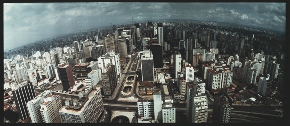 Descrição da imagem: Fotografia panorâmica aérea colorida da cidade de São Paulo, com muitos prédios altos, capturada de cima. A imagem exibe um horizonte recurvado de uma vasta área urbana, a partir da área da Avenida Paulista, com ruas e avenidas que se cruzam, e prédios de diferentes tamanhos e estilos arquitetônicos. O céu está nublado. Fim da descrição da imagem.