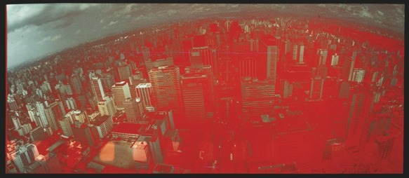 Descrição da imagem: Fotografia panorâmica aérea colorida da cidade de São Paulo, com muitos prédios altos, capturada de cima, com um filtro avermelhado aplicado sobre toda a imagem. Os prédios e estruturas urbanas são visíveis. Fim da descrição da imagem.