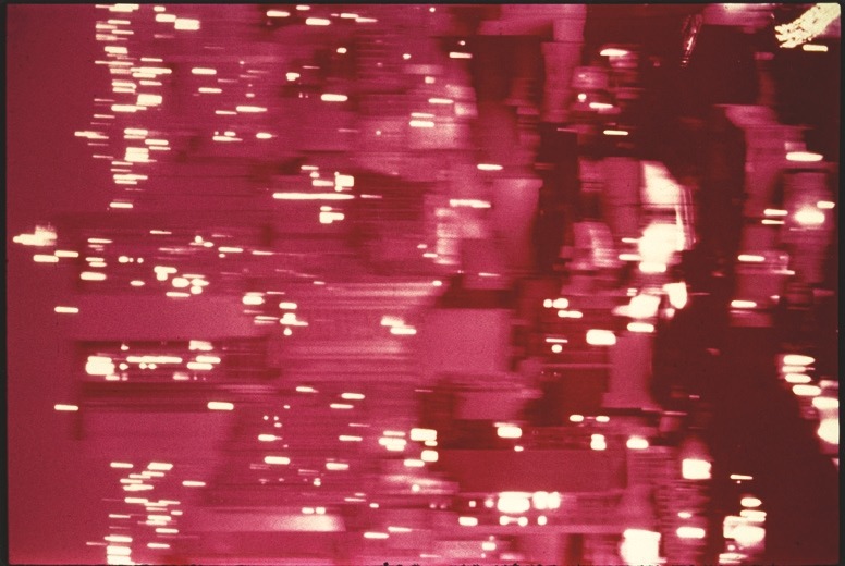 Descrição da imagem: Fotografia horizontal colorida com uma forte tonalidade avermelhada e abstrata. A imagem parece mostrar uma paisagem urbana à noite, com luzes de edifícios e janelas borradas e distorcidas. Fim da descrição da imagem.