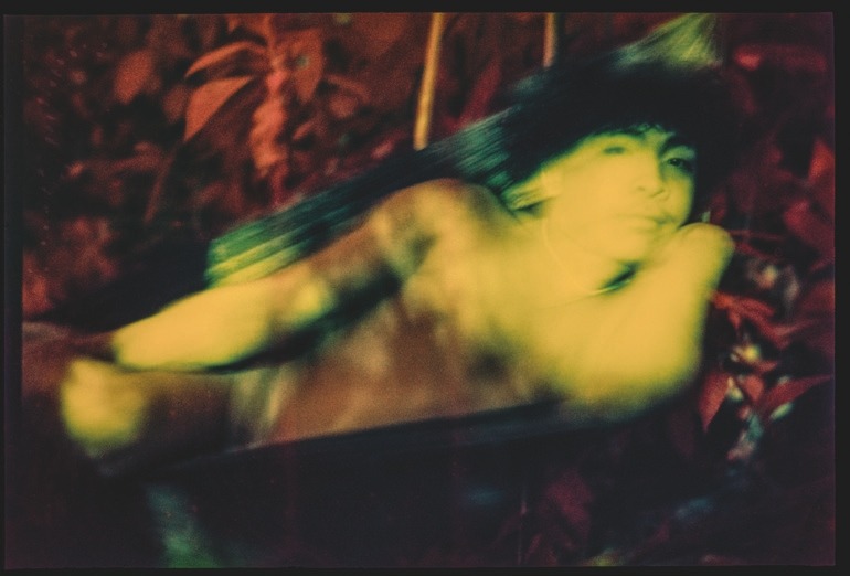 Descrição da imagem: Fotografia horizontal colorida de um jovem indígena yanomami deitado em uma rede. Está cercada por vegetação em tons de vermelho e laranja, com expressão calma. Seu corpo está iluminado com luz amarela esverdeada. Fim da descrição da imagem.