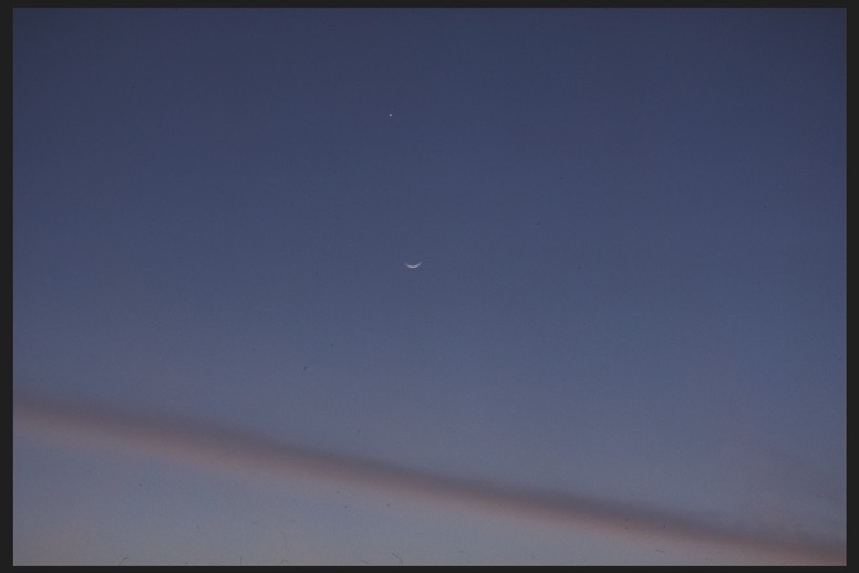 Descrição da imagem: Fotografia horizontal colorida de um céu noturno, com um fundo azul escuro. No centro da imagem, uma fina lua crescente é visível, com um ponto brilhante logo acima. Uma linha fina de nuvens cruza a parte inferior da imagem. Fim da descrição da imagem.