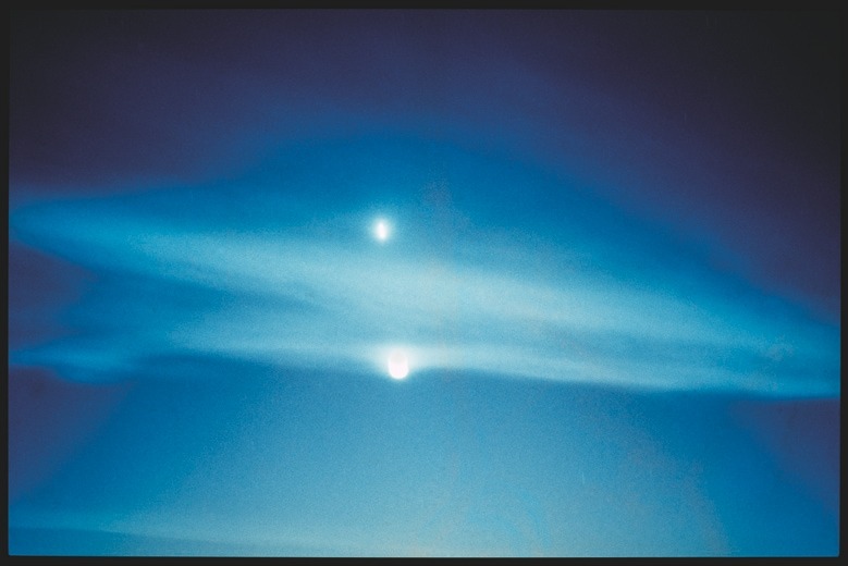 Descrição da imagem: Fotografia horizontal colorida de um céu azul com um efeito de difusão de luz. Dois pontos brancos e brilhantes, estão no centro da imagem, flutuando sobre uma camada fina de nuvens. O efeito visual cria uma sensação etérea, com um gradiente suave de tons de azul. Fim da descrição da imagem.