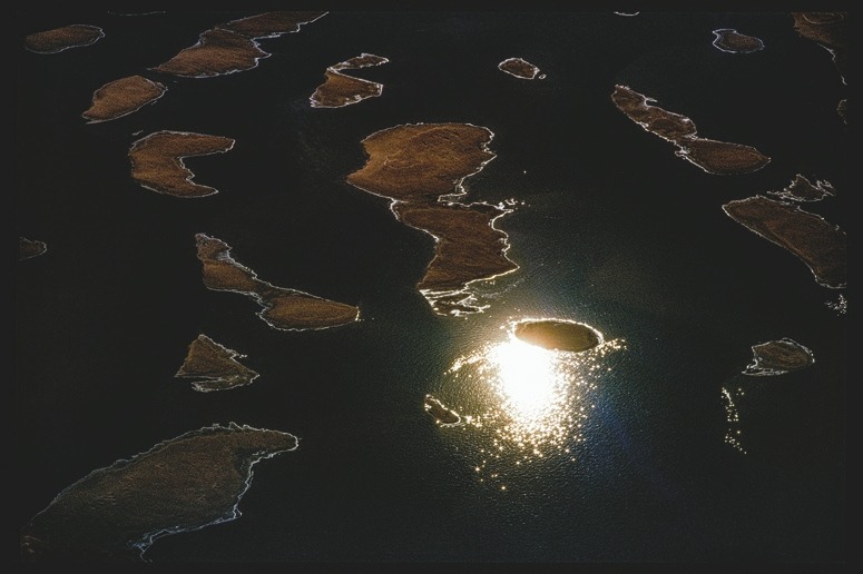 Descrição da imagem: Fotografia horizontal colorida de várias pequenas ilhas dispersas em um corpo d'água de um rio. A luz do sol reflete na água, criando um brilho intenso em uma das áreas próximas às ilhas. As ilhas têm bordas irregulares e estão cercadas por água escura e reflexos luminosos. Fim da descrição da imagem.