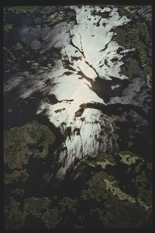 Descrição da imagem: Fotografia horizontal colorida de uma paisagem natural vista de cima, com o reflexo do sol iluminando um grande corpo d'água de rio. A luz reflete de forma difusa, criando formas brilhantes na superfície da água, cercadas por áreas verdes e escuras de vegetação. Fim da descrição da imagem.