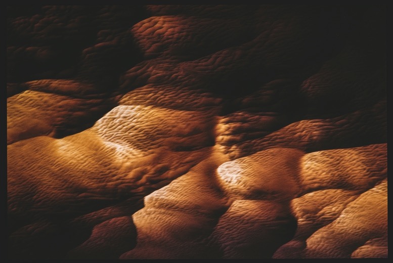 Descrição da imagem: Fotografia colorida horizontal. A imagem apresenta uma superfície texturizada com padrões ondulados e irregulares, iluminada por uma luz que projeta diferentes tonalidades de marrom e laranja, criando áreas de sombra e destaque. As ondulações parecem lembrar uma formação de dunas, vista de cima, com variações suaves nas formas e relevo. Fim da descrição da imagem.