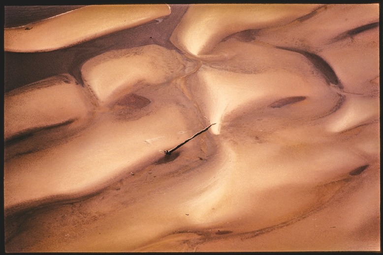 Descrição da imagem: Fotografia colorida horizontal. A imagem mostra uma superfície arenosa com cores que variam entre tons claros de bege e marrom. Há formas suaves e curvas que parecem ser resultado de sedimentos moldados por água ou vento, com um rasgo diagonal escuro que corta a superfície. A textura lembra uma formação de areia, vista de cima. Fim da descrição da imagem.