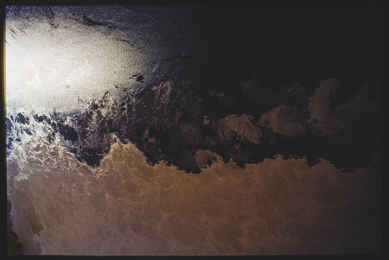 Descrição da imagem: Fotografia colorida horizontal. A imagem exibe um corpo de água onde há uma transição de cores, com uma porção escura no topo da imagem e uma área mais clara e marrom na parte inferior. A superfície da água reflete parcialmente a luz, criando uma divisão clara entre as áreas escuras e claras. A textura sugere movimento nas águas no encontro de dois rios, como entre o Rio Negro e o Solimões. Fim da descrição da imagem.
