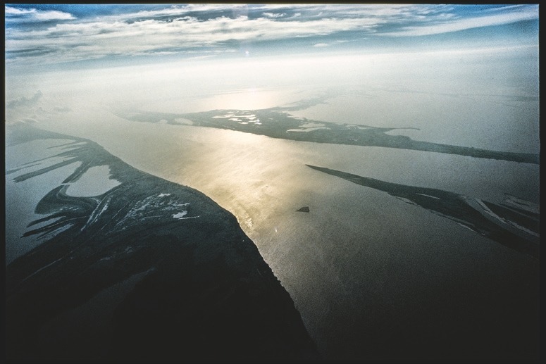 Descrição da imagem: Fotografia colorida horizontal e aérea do rio Negro Anavilhanas. A imagem mostra uma grande extensão de água, com a luz do sol refletida em sua superfície, formando um brilho dourado ao longo do rio. A costa à esquerda está delineada por faixas de terra e vegetação que se projetam em direção à água. O céu ao fundo é parcialmente nublado, com tons suaves de azul e branco. Fim da descrição da imagem.