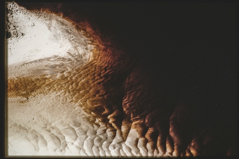 Descrição da imagem: Fotografia colorida horizontal. A imagem exibe uma superfície de encontro de águas de rios distintos que se assemelha a uma superfície arenosa, com variação de cores que vai do branco ao marrom escuro. A textura da das águas é ondulada em sua parte central e mais espumada à esquerda da imagem. À direita, há uma grande porção escura, que parece ser sombra, criando um forte contraste com as áreas mais claras à esquerda. Fim da descrição da imagem.
