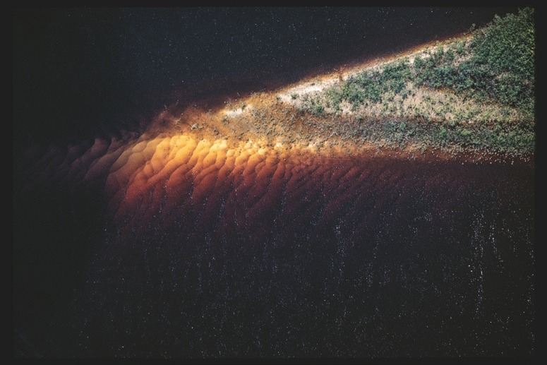 Descrição da imagem: Fotografia colorida horizontal. A imagem mostra uma porção de terra triangular cercada por água escura. A forma é de um triângulo retângulo bem alongado em sua base, e está no canto superiro direito da imagem. A luz do sol ilumina parcialmente a terra, realçando os tons alaranjados da areia, enquanto a vegetação verde densa cobre o topo do triângulo. O contraste entre a água escura e a terra iluminada destaca o formato triangular da porção de terra. Fim da descrição da imagem.