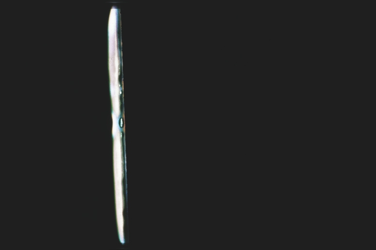 Descrição da imagem: Fotografia colorida horizontal. A imagem exibe uma forma alongada e vertical de um objeto estreito e brilhante, contra um fundo totalmente preto. O objeto tem tons perolados, refletindo uma leve variação de cores, parecendo um feixe de luz ou um reflexo em uma superfície líquida. Fim da descrição da imagem.