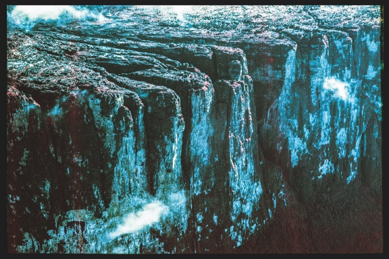 Descrição da imagem: Fotografia colorida vertical. A imagem retrata uma impressionante formação de falésias altas, com suas paredes verticais recortadas, iluminadas por tons azulados e avermelhados . A superfície superior das falésias é coberta por vegetação, enquanto nuvens baixas e sombras intensificam o contraste entre o topo e a face rochosa. As cores foram obitidas por meio de manipualção na revelação da imagem. Fim da descrição da imagem.