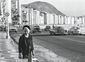 Descrição da imagem: Fotografia em preto e branco. Em primeiro plano, à esquerda, uma mulher idosa em uma calçada, vista de frente, usa uma boina com o cabelo curto aparente no topo da testa, colar, casaco e saia. Com o braço esquerdo, segura o que sugere ser uma bolsa. Ao fundo, muitos prédios contornam a orla de uma praia, e uma extensa avenida com alguns carros Fim da descrição da imagem.