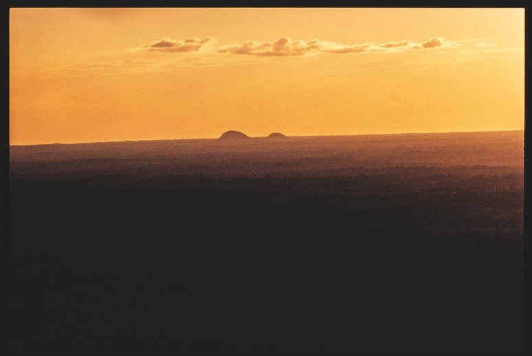 Descrição da imagem: Fotografia colorida horizontal. A imagem retrata um pôr do sol, com o céu em tons de laranja e amarelo. No horizonte, duas elevações arredondadas se destacam, enquanto a parte inferior da imagem mostra uma vasta área escura de vegetação e relevo plano, contrastando com a luz intensa do céu. Fim da descrição da imagem.