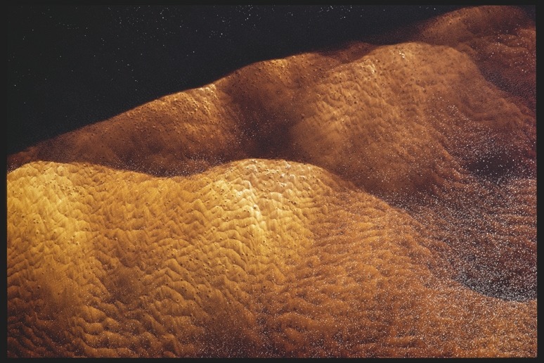 Descrição da imagem: Fotografia colorida horizontal. A imagem apresenta uma superfície ondulada de areia ou sedimentos sob uma camada de água de um rio, em tons de marrom claro e dourado, com textura semelhante a pequenas dunas. A iluminação cria sombras suaves que realçam o relevo das ondulações. O fundo escuro contrasta com as formas claras em primeiro plano. Fim da descrição da imagem.