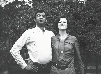 Descrição da imagem: Fotografia em preto e branco de George Love, à esquerda, e Claudia Andujar, à direita, em pé. Ele usa camisa de manga comprida clara e calça escura, sorri e está com a mão esquerda na cintura e a direita abraça Claudia pela cintura. Ela usa blusa de manga comprida escura, calça escura e um lenço amarrado no pescoço e abraça George pela cintura com a mão esquerda. Ao fundo, algumas árvores. Fim da descrição da imagem.
