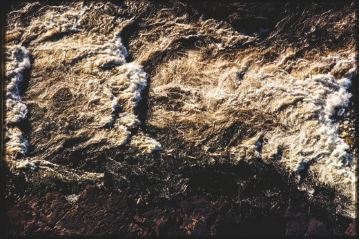 Descrição da imagem: Fotografia colorida vertical. A imagem mostra um corpo de água com corredeiras turbulentas, onde a espuma branca das ondas contrasta com as águas escuras. As correntes de água formam padrões caóticos, criando uma textura intensa de movimento. Nas lateriais da imagem, observa-se as margens pedregosas do rio. Fim da descrição da imagem.