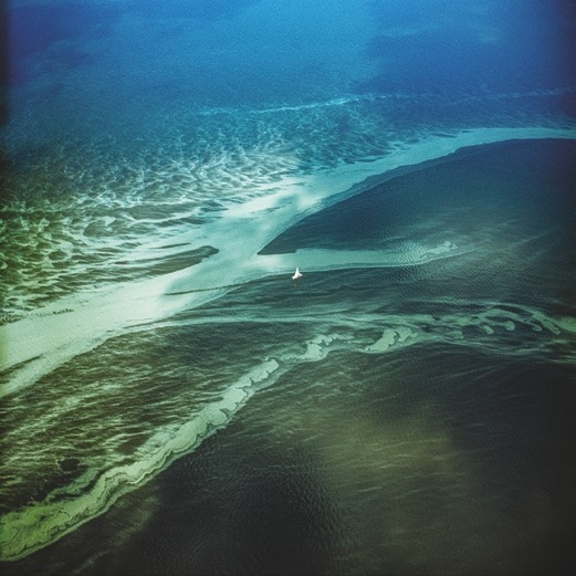 Descrição da imagem: Fotografia colorida quadrada. A imagem retrata uma vista aérea de um delta, onde bancos de areia e canais de água se misturam. A água, em tons de azul e verde, forma padrões fluidos, enquanto um barco branco aparece no centro, destacando-se no vasto cenário. A imagem tem tonalidades azuis, verdes e amarelas. Fim da descrição da imagem.