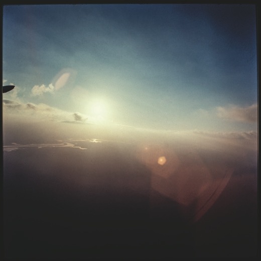 Descrição da imagem: Fotografia colorida quadrada. A imagem apresenta um pôr do sol visto do alto, na perspectiva de um avião, com o céu em tons suaves de azul e laranja. As nuvens dispersas adicionam profundidade à cena, enquanto a luz do sol se reflete nas águas ao longe. O horizonte aparece embaçado devido à leve névoa. Fim da descrição da imagem.