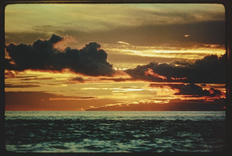 Descrição da imagem: Fotografia colorida horizontal. A imagem mostra um pôr do sol sobre um rio amazônico, com um corpo d'água imenso, e o céu em tons de laranja, amarelo e vermelho. Nuvens escuras cortam o céu, criando um contraste com as cores vibrantes ao fundo. O rio, em primeiro plano, reflete suavemente a luz do céu. Fim da descrição da imagem.