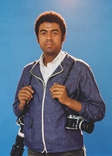 Descrição da imagem: fotografia colorida de George Love, em pé. Ele usa jaqueta jeans azul sobre camisa branca com listras na vertical, e segura duas câmeras fotográficas pretas penduradas por alças pretas, uma em cada ombro. As câmeras são pretas e tem grandes lentes. A parte de cima de seu corpo está iluminada por uma luz que vem de cima. O fundo é todo azul. Fim da descrição da imagem.