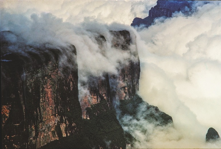 Descrição da imagem: Fotografia colorida horizontal. A imagem mostra uma cadeia de montanhas, cobertas por nuvens espessas, com picos rochosos emergindo das nuvens. A luz suave ilumina as faces rochosas. Fim da descrição da imagem.