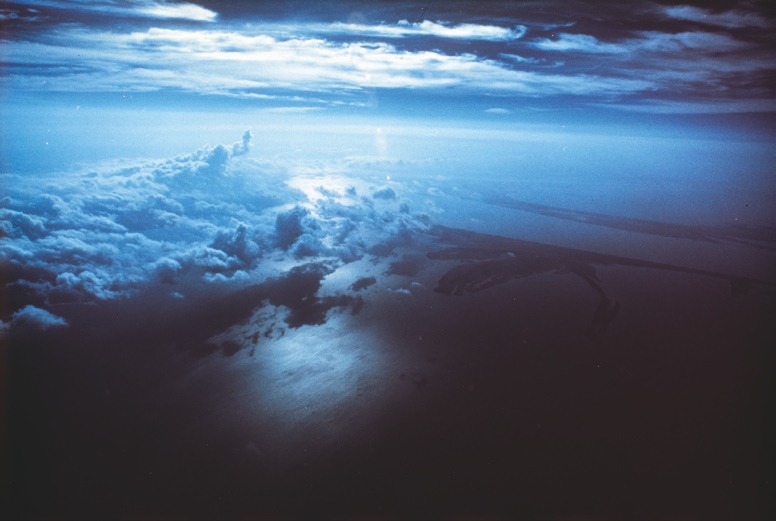 Descrição da imagem: Fotografia colorida horizontal. A imagem retrata uma vista aérea de um grande corpo d'água e formações de nuvens densas ao longo do horizonte. A luz do sol se reflete suavemente na água, em um tom azulado que permeia toda a cena, destacando a imensidão da paisagem. Fim da descrição da imagem.