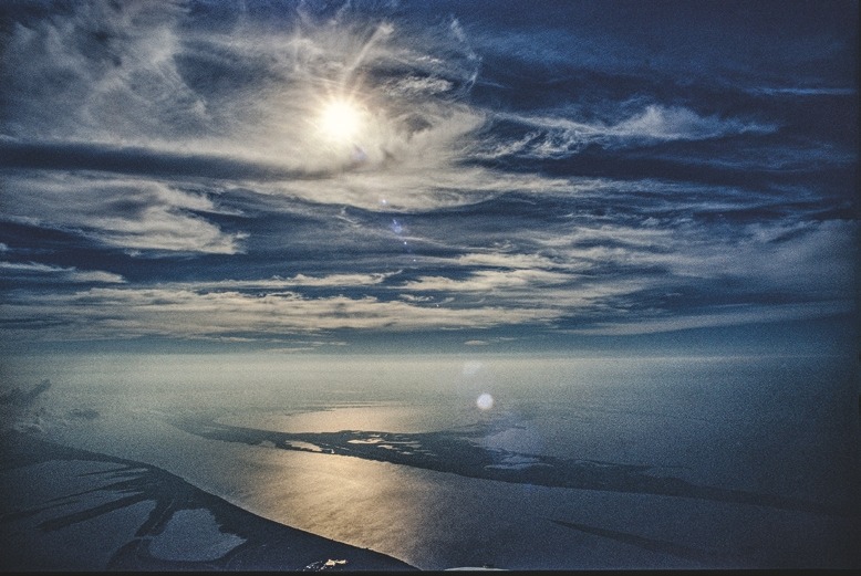 Descrição da imagem: Fotografia colorida horizontal. A imagem mostra uma vista aérea de um rio serpenteando através de uma paisagem parcialmente iluminada pelo sol. O céu está coberto por nuvens finas, com o sol brilhando intensamente, criando reflexos na superfície da água. Fim da descrição da imagem.