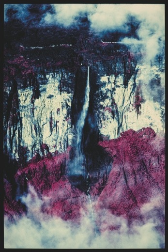 Descrição da imagem: Fotografia colorida vertical. A imagem exibe uma cachoeira alta caindo de um penhasco rochoso cercado por neblina e nuvens. O penhasco tem tons claros de bege e branco, enquanto o solo e a vegetação ao redor aparece em tons avermelhados. A água da cachoeira se destaca contra as cores das rochas. Fim da descrição da imagem.