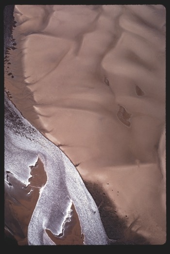 Descrição da imagem: Fotografia colorida vertical. A imagem mostra uma área de areia lisa, com leves ondulações na superfície, em tons de marrom claro. À esquerda, uma parte da areia é atravessada por um curso de rio de águas escuras e com um brilho intenso branco do reflexo do sol, criando um contraste de texturas e cores. Fim da descrição da imagem.