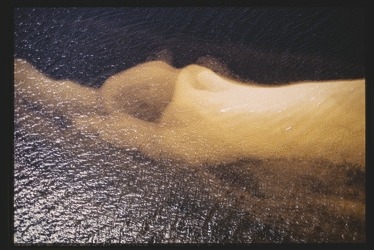 Descrição da imagem: Fotografia colorida horizontal. A imagem mostra um banco de areia sob um corpo d'água iluminado por luz, com a areia clara contrastando com a água escura, mais profunda, ao redor. A forma da areia cria uma curva suave em direção à água, enquanto o movimento da água ao redor forma leves ondulações na superfície. Fim da descrição da imagem.