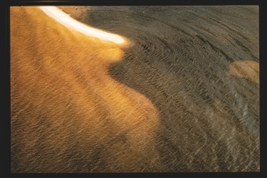 Descrição da imagem: Fotografia colorida horizontal. A imagem apresenta uma área de água e areia, com uma parte da areia iluminada em um tom dourado. A transição entre a areia clara e a área escura forma uma curva suave, sugerindo uma interação entre água e sedimento, com parte da areia sob a água e outra parte acima dela. Fim da descrição da imagem.