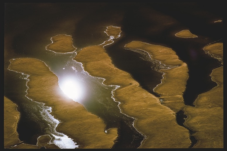 Descrição da imagem: Fotografia colorida horizontal. A imagem captura uma série de ilhas ou bancos de areia iluminados, cercados por água escura. A luz reflete intensamente na água, criando um ponto de brilho no centro da imagem, enquanto os bancos de areia formam formas irregulares que se estendem pela cena. Fim da descrição da imagem.