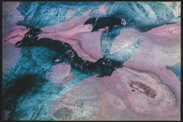 Descrição da imagem: Fotografia aérea colorida horizontal mostrando uma superfície com padrões abstratos em tons de azul e rosa. Manchas escuras e irregulares atravessam a imagem, destacando-se sobre o fundo texturizado. As cores formam formas orgânicas, lembrando traços de tinta misturada em um líquido. Fim da descrição da imagem.