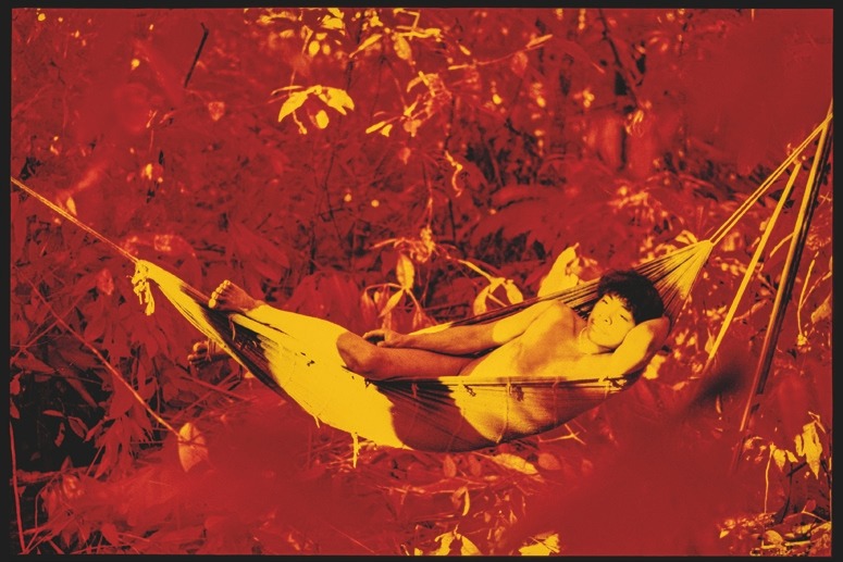 Descrição da imagem: Fotografia horizontal colorida com cores manipuladas de uma pessoa deitada em uma rede. A pessoa, um jovem indígena Yanomami, está relaxado, com um braço apoiado na cabeça e o pé direito saindo sobre a rede. O fundo da imagem é dominado por uma vegetação em tons intensos de vermelho e laranja. Fim da descrição da imagem.