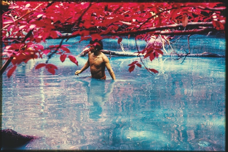 Descrição da imagem: Fotografia horizontal com cores manipuladas de um jovem Yanomami, com o tronco nu, pintado com manchas arrendodas e usando um pequeno colar de missangas, está submerso até a cintura em um rio de águas azul-claras. Ramos com folhas vermelhas cobrem parcialmente a parte superior da imagem, criando um forte contraste com a cor da água. Fim da descrição da imagem.