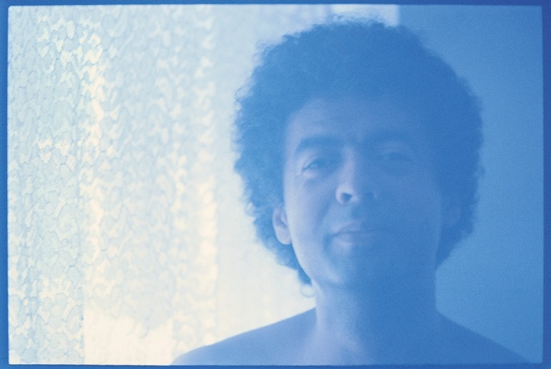 Descrição da imagem: Fotografia horizontal colorida do torso de George Love. A imagem está levemente desfocada, e ele está com expressão suave e um leve sorriso, ao lado de uma janela com cortinas rendadas claras, que permitem a entrada de uma luz suave. Fim da descrição da imagem.