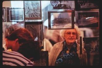 Descrição da imagem: Imagem colorida e horizontal. Em um ambiente interno, uma mulher idosa de cabelos brancos e óculos está em pé, olhando para várias fotografias em preto e branco expostas em frente a ela. À esquerda, parcialmente visíveis, estão duas outras pessoas, uma de cabelo castanho e outra de cabelo escuro, observando as fotos. As fotografias estão penduradas em uma estrutura de vidro ou acrílico, criando reflexos e transparências, podendo ver tanto as imagens quanto o que há por detrás delas.Fim da descrição da imagem.