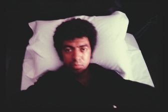 Descrição da Imagem: Fotografia de George Love deitado em uma cama. Ele tem cabelo afro volumoso, uma camisa escura e está deitado de costas, olhando para a câmera. Ele está em um quarto escuro com um travesseiro branco sob a cabeça. A foto está um pouco embaçada. Fim da descrição da imagem.