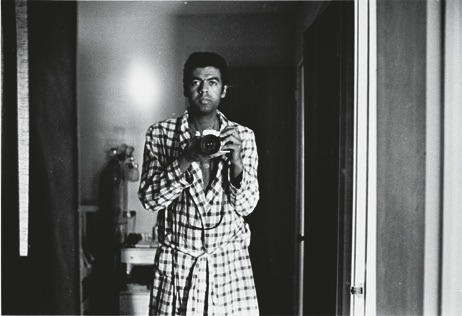 Descrição da imagem: fotografia em preto e branco de George Love se olhando no espelho. Ele tem pele negra, cabelo curto e escuro, usa um roupão xadrez e segura uma câmera fotográfica apontada para o espelho. No reflexo, vemos um cômodo com objetos difusos. Fim da descrição da imagem.