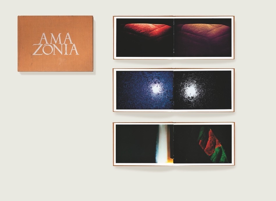 Descrição da imagem: Imagem colorida e horizontal. Montagem do livro "AMAZÔNIA" aberto em três páginas distintas e fechado, mostrando a parte de trás do livro. Na montagem, na parte esquerda superior está uma imagem de alto contraste preto e branca. Ela mostra uma mistura de um rio espumado com o reflexo do céu. A segunda imagem mostra um céu azul profundo com dois pontos luminosos entre uma macha branca de nuvem. A terceiro imagem apresenta uma vista aérea de ondas no mar, com tons de roxo e azul. Ao lado direito, há o livro fechado com capa marrom e uma pequena imagem colorida no centro. Está imagem é de uma pessoa indigena deitada em uma rede. Fim da descrição da imagem.